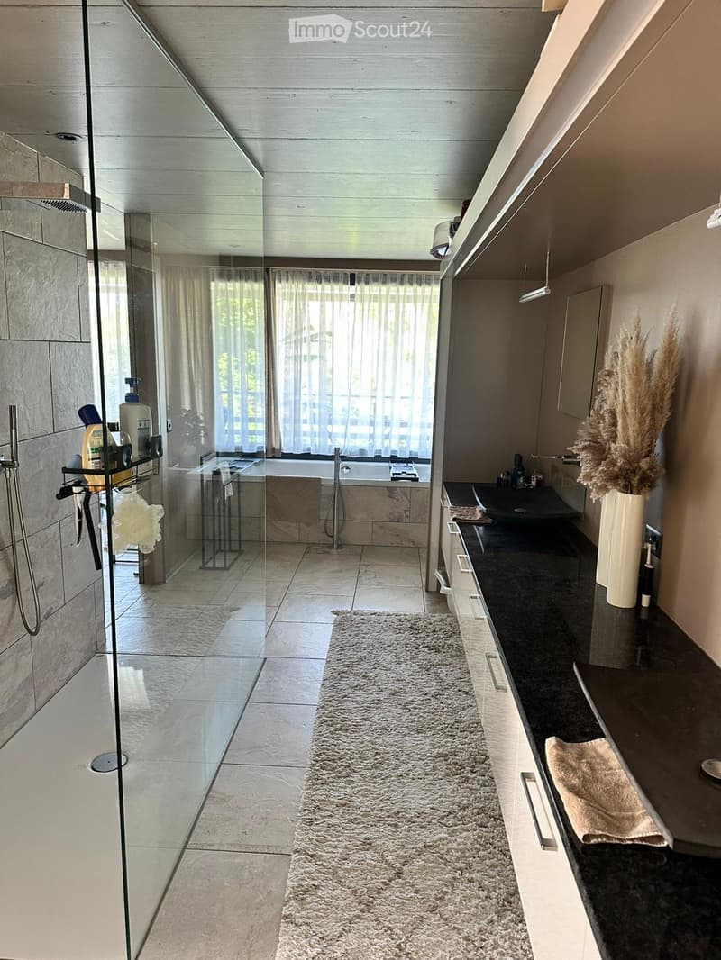 Modern bathroom with a bathtub, glass-enclosed shower, sink, and vanity. Features a beige tiled floor, a plush rug, and decorative vase with dried plants.