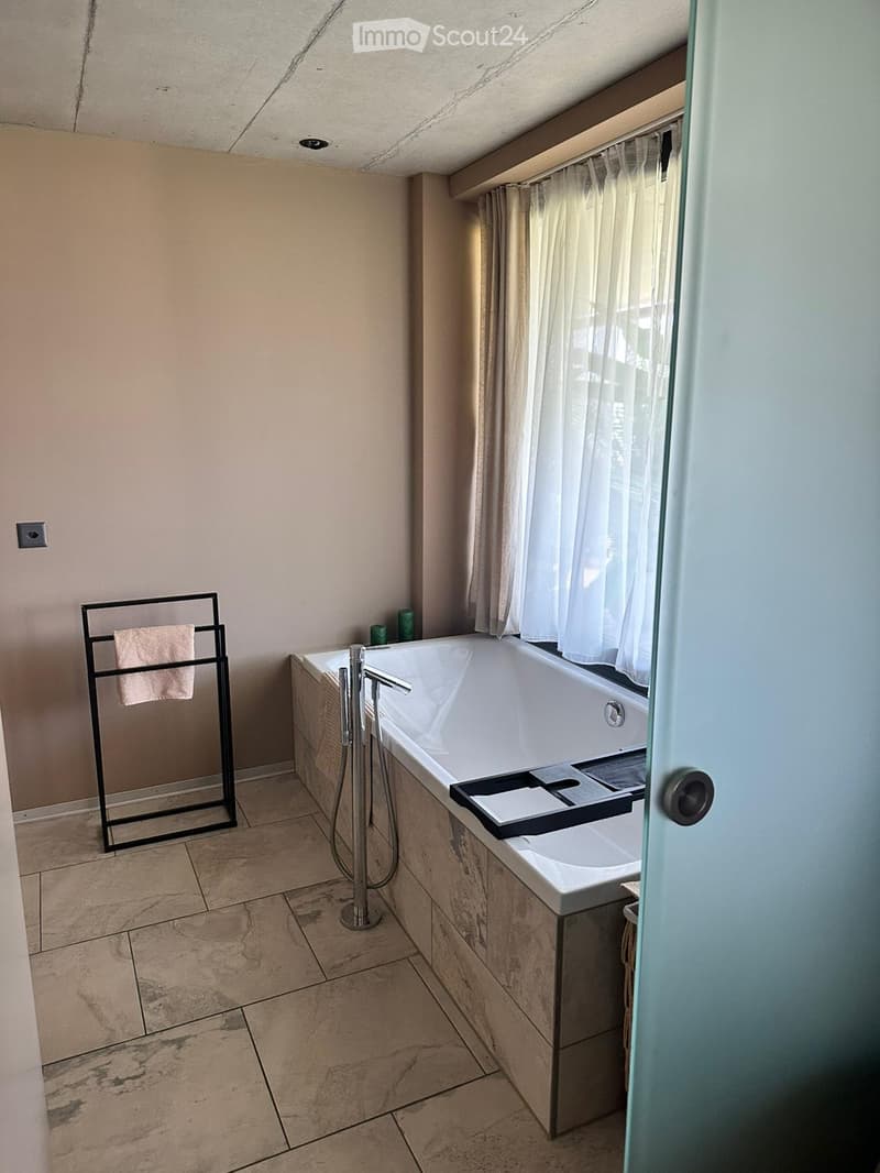 Modern bathroom with a large bathtub, sleek faucet, towel rack with a towel, and a window with white curtains.
