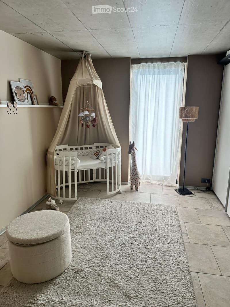 A well-lit, modern nursery with a white crib under a beige canopy, a plush giraffe toy, a round beige stool, a large shaggy rug, and a floor lamp near the window.
