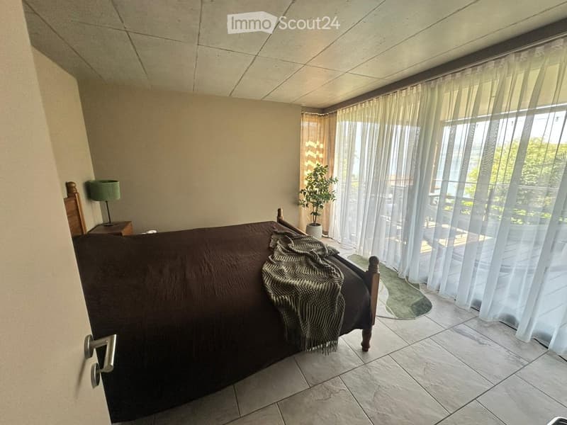 Cozy bedroom with a large bed, nightstand, lamp, and a potted plant near the window. The room features tiled flooring and large windows with sheer curtains.