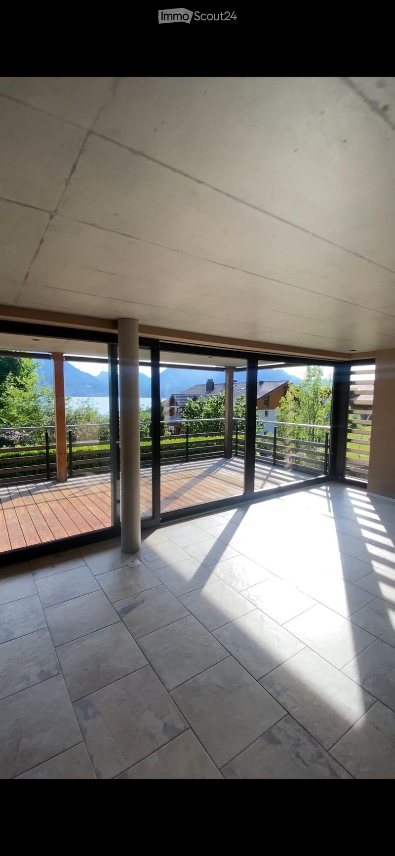 Bright, empty room with large glass sliding doors leading to a wooden deck, offering a scenic mountain and lake view.