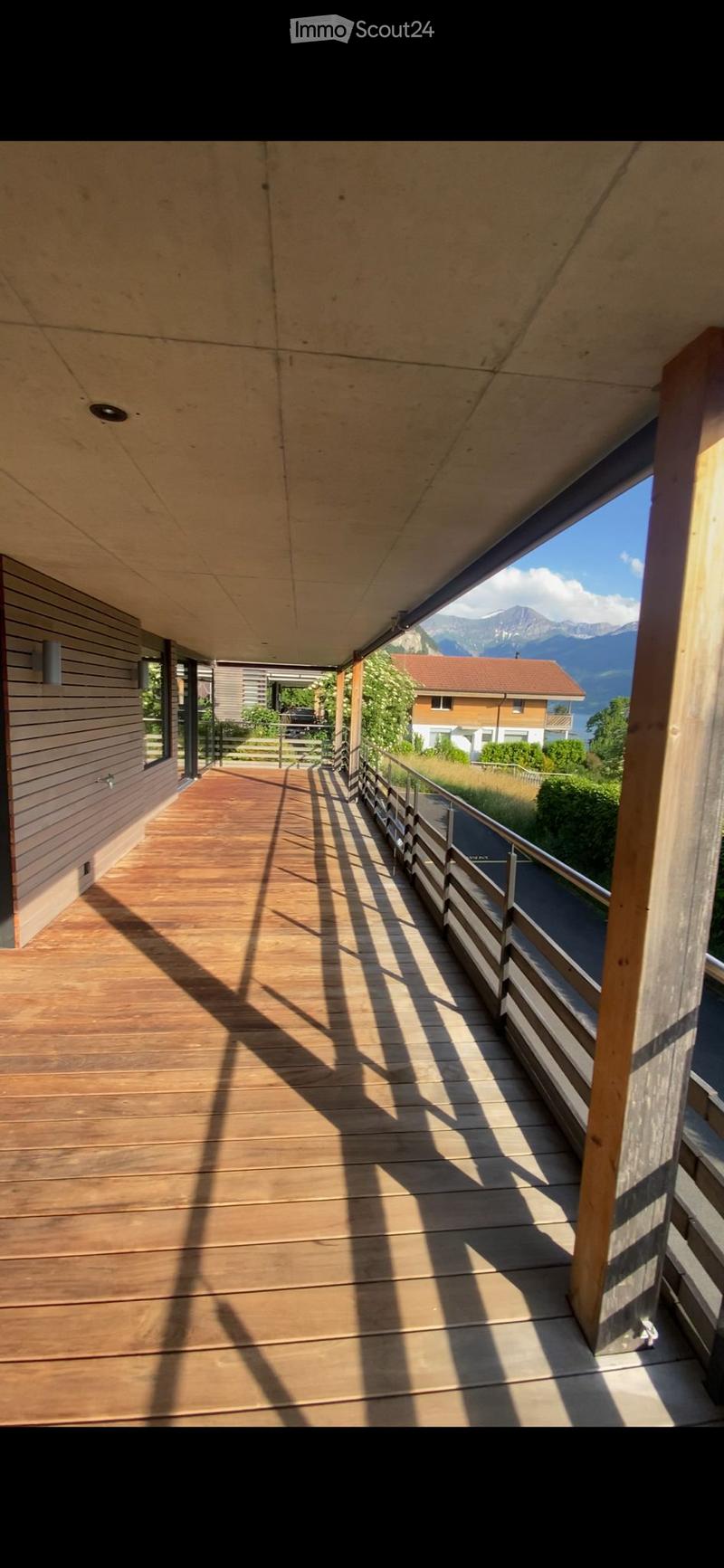 Wooden balcony with a railing, overlooking a scenic mountain view and surrounding houses.