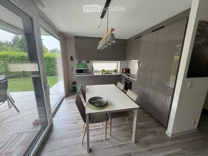 Modern kitchen with grey cabinets, a white dining table with four chairs, and a microwave on the counter. Large windows provide natural light and a view of the garden.