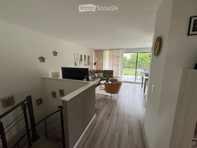 Modern living room with wooden flooring, a sectional sofa, coffee table, armchair, and a television. The room features large windows with sheer curtains and a view of a garden, with additional seating and a small table visible through the glass door.