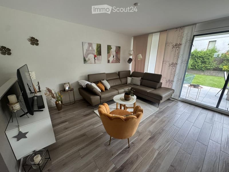 Modern living room with a grey sectional sofa, a round coffee table, a yellow armchair, a TV, and decorative items including vases and candles. Large windows with sheer curtains lead to a patio area.
