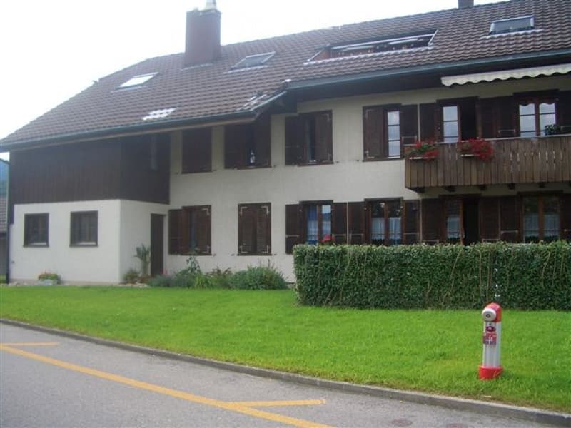 Two-story house with a red-tiled roof, white walls, and multiple windows with shutters. Features a well-maintained lawn and a small garden area.