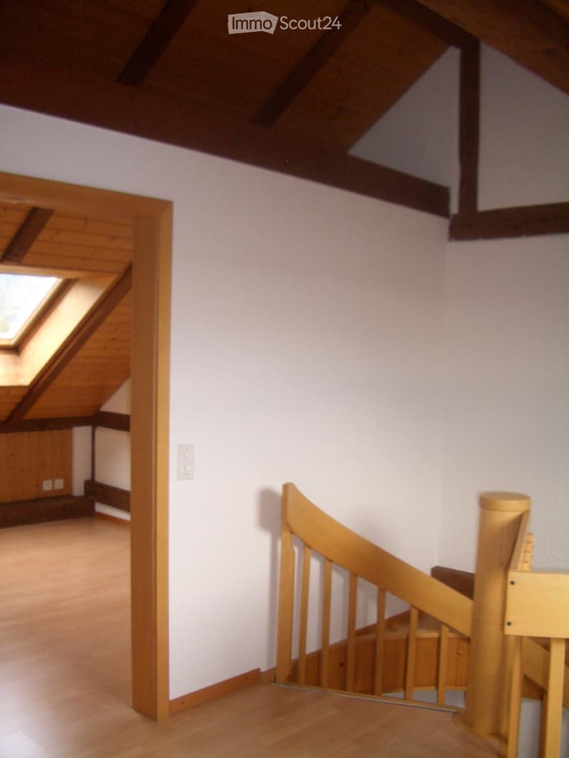 Wooden staircase with a handrail and open wooden balustrade, leading to an attic space with exposed wooden beams and a skylight window.