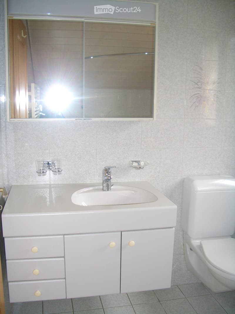 Modern bathroom with a white sink, faucet, and toilet. The sink has a built-in cabinet with drawers and doors. A mirror is mounted above the sink, and two cups are placed on the sink counter.