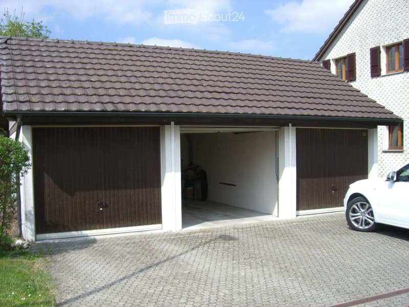 Two-story house with a tiled roof, white exterior, and a large garage with wooden doors. A white car is parked in front of the garage.