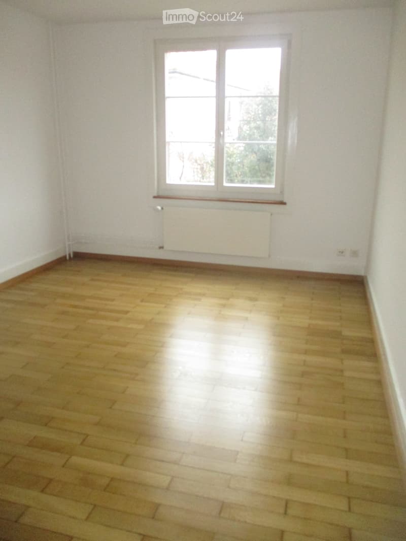 Empty room with wooden flooring, white walls, and a large window with a view of greenery outside.