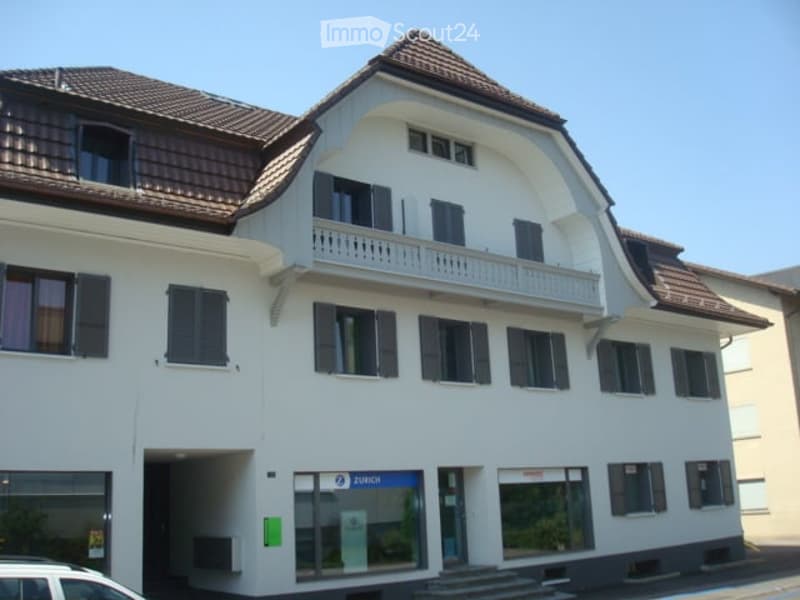 Building. A two-story white building with a brown tiled roof, multiple windows with shutters, and a small balcony on the upper floor. The ground floor features a glass entrance with a sign that reads "ZURICH".