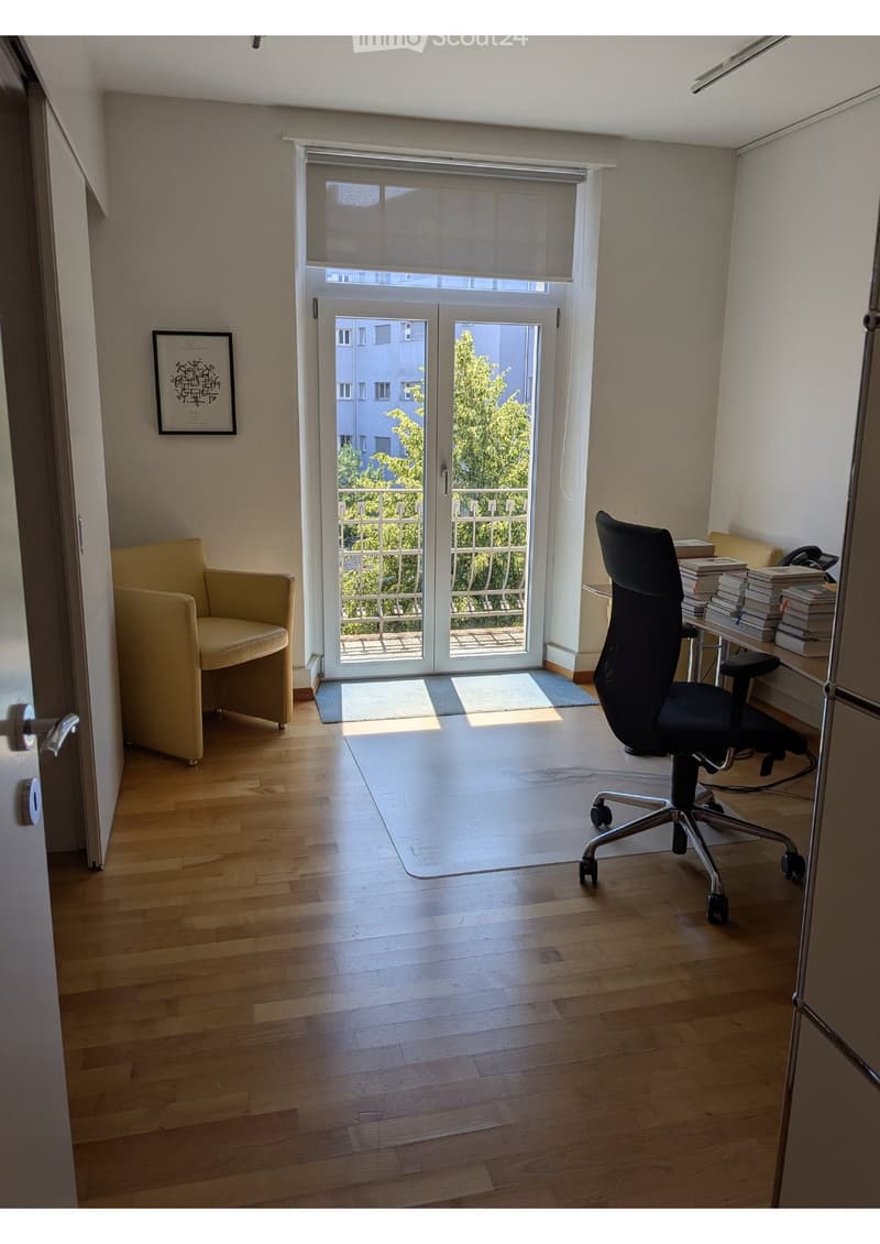 Bright room with wooden flooring, large sliding glass door leading to a balcony, a beige armchair, an office chair, a small table with books and a telephone, and a picture frame on the wall.