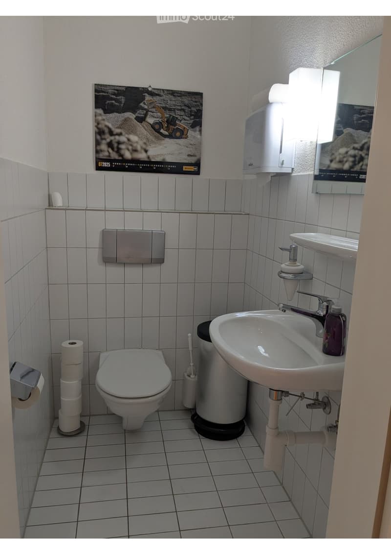 A small bathroom with a toilet, sink, mirror, and a stack of toilet paper rolls. The walls are tiled, and a poster is visible on one wall.