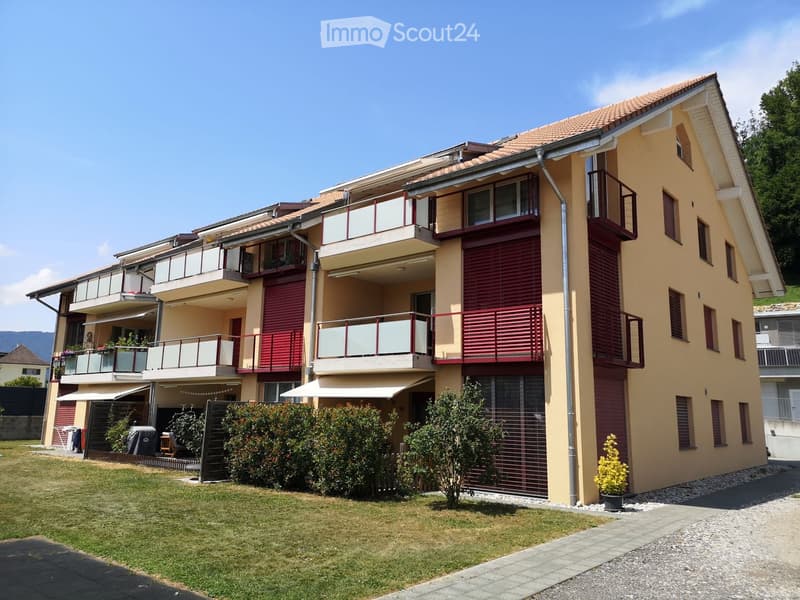 Modern two-story residential building with beige and red exterior, multiple balconies, and a well-maintained lawn in front.