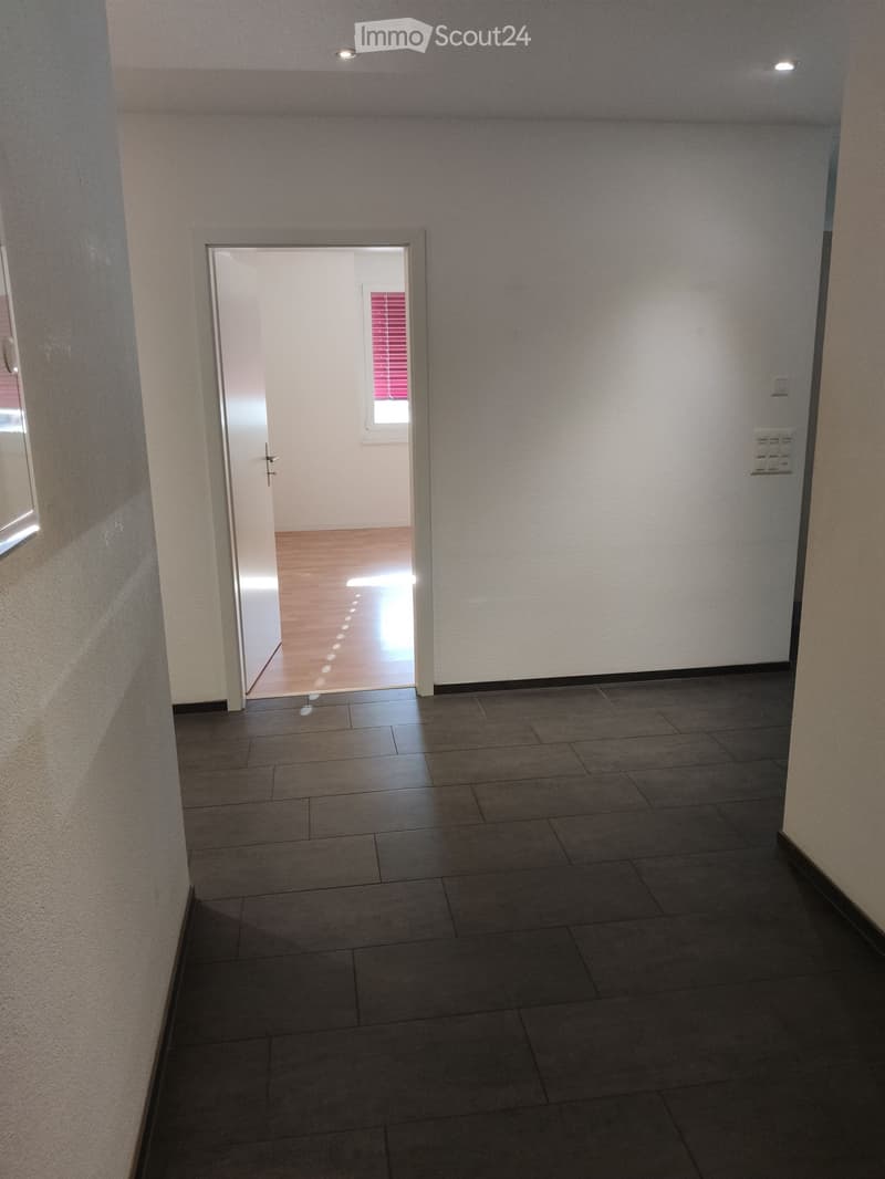 Empty hallway with tiled floor and light-colored walls, featuring a door leading to another room with wooden flooring and a window with blinds.
