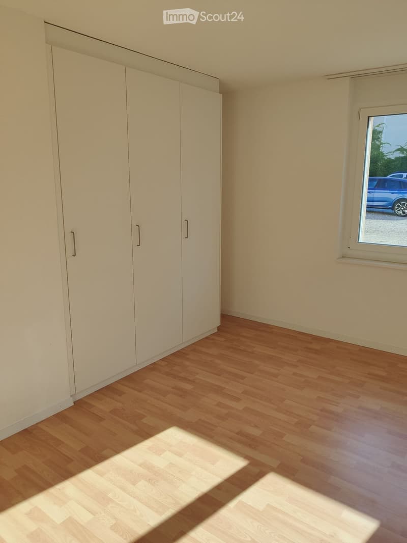 Empty room with light wooden flooring, white walls, and built-in white wardrobes. A window on the right side shows a view of a parked car outside.