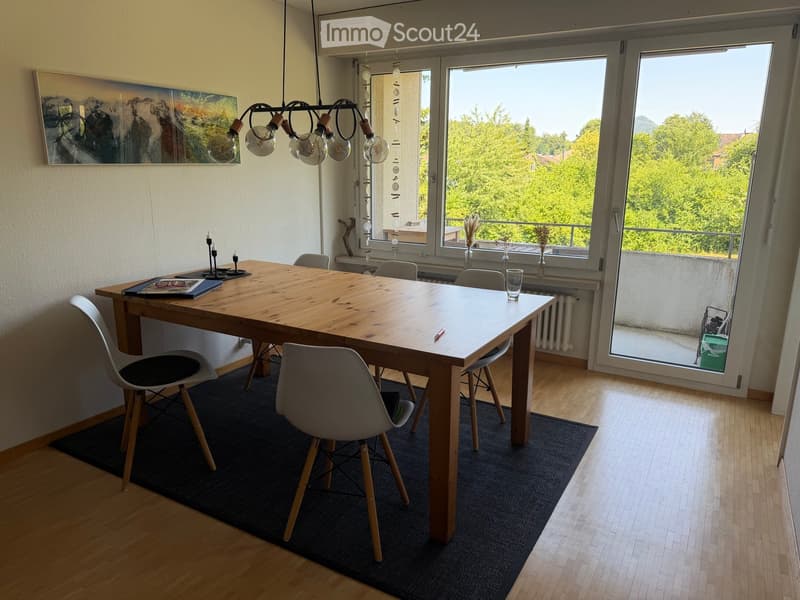 Dining room with a wooden table, chairs, a painting on the wall, and a hanging lamp. Large windows offer a view of greenery outside.