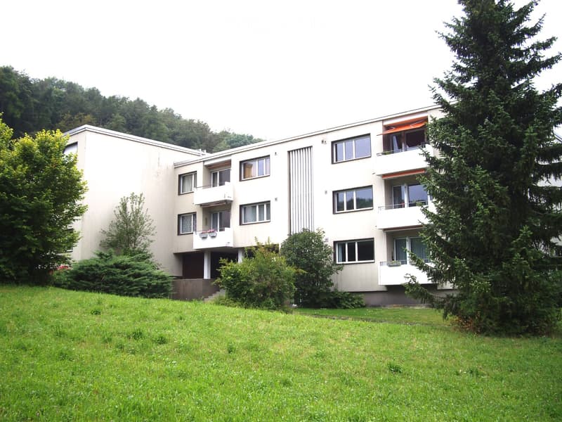 Modern multi-story residential building with balconies, surrounded by greenery and trees.