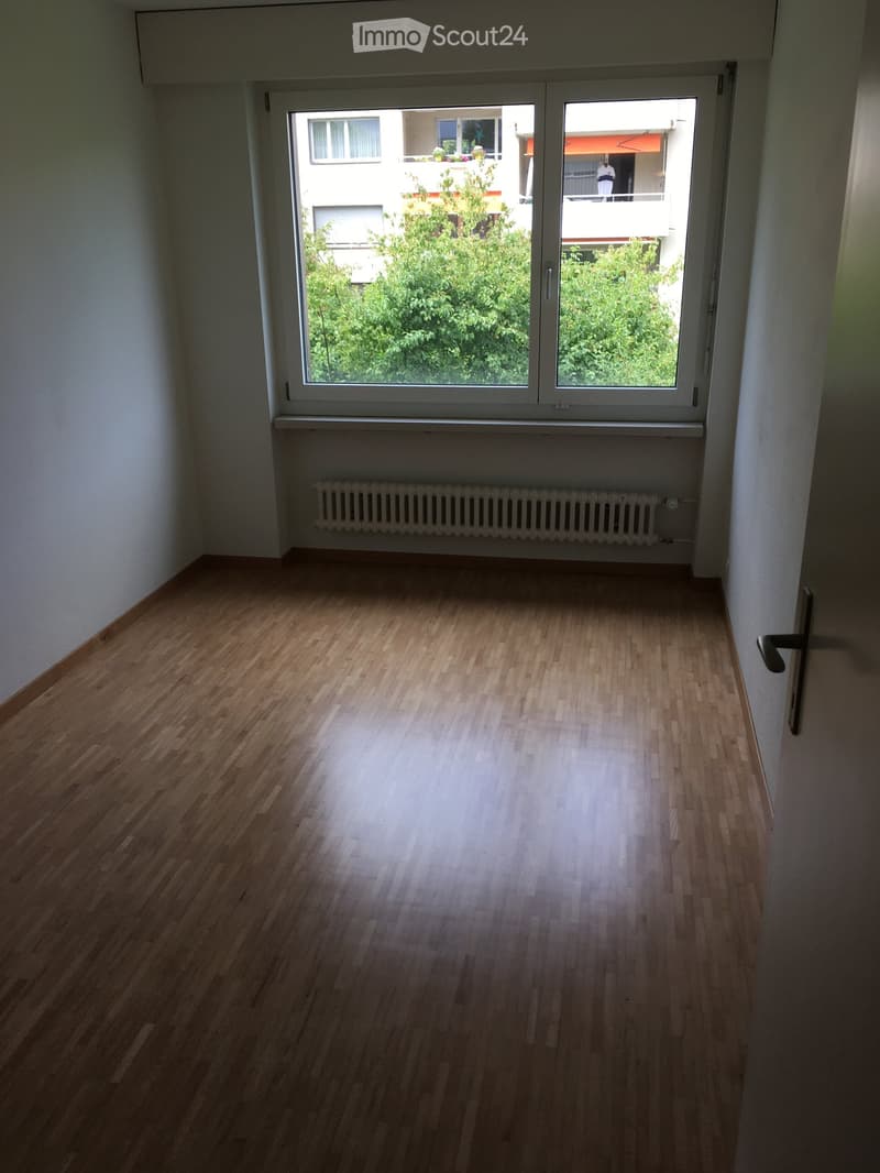 Empty room with wooden flooring, white walls, large window with view of greenery, radiator below window, and door on right side.
