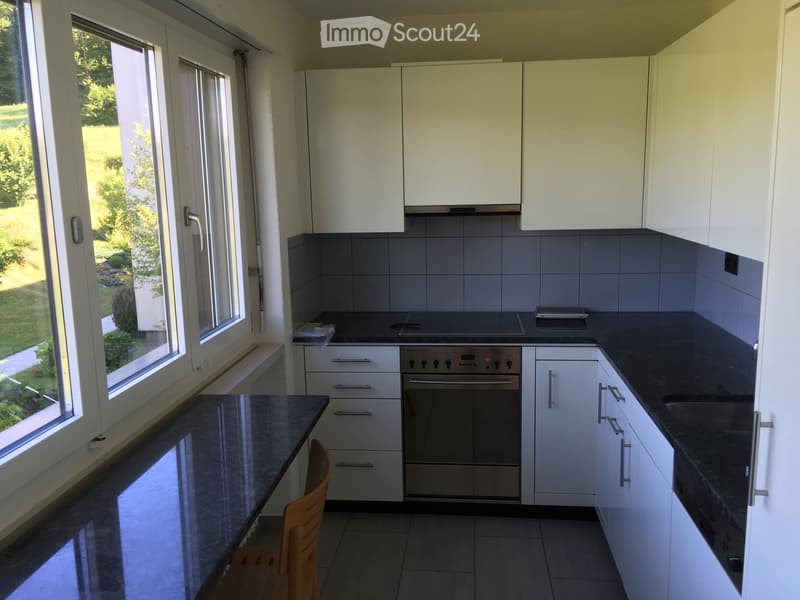 Modern kitchen with white cabinets, dark granite countertops, built-in oven, and a wooden chair near the island.