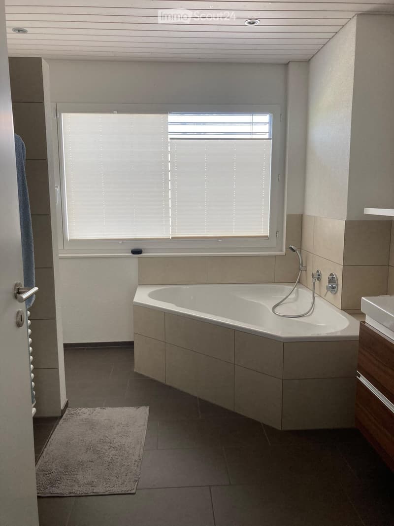 Modern bathroom with a corner bathtub, a sink with a wooden cabinet, a window with blinds, and a small rug on the floor.