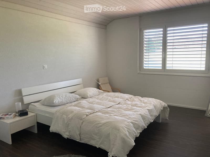 A modern bedroom with a large bed, white bedding, a nightstand with a lamp and book, a wooden chair, and large windows with blinds.