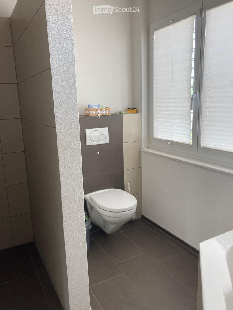 Modern bathroom with a toilet, sink, and a wicker basket on the wall. The room features tiled walls and a window with blinds.