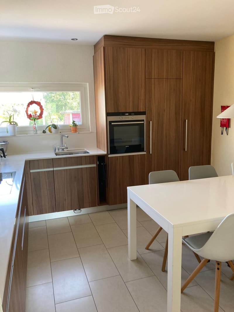 Modern kitchen with wooden cabinets, stainless steel oven, white countertop, sink, and a small dining area with a white table and chairs.
