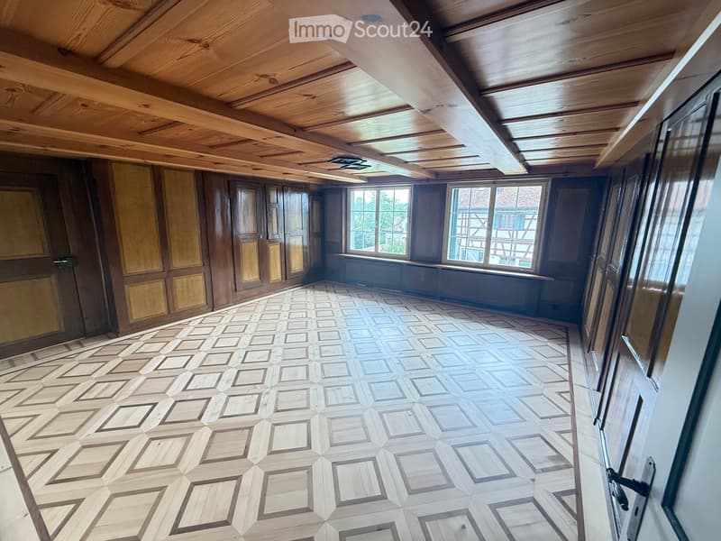 Empty room with wooden walls, geometric patterned wooden floor, large windows, and exposed wooden ceiling beams.