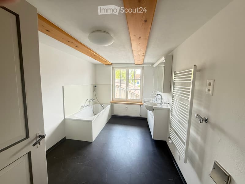 Modern bathroom with a bathtub, double sink, mirror, radiator, and large window offering a view of the roof.