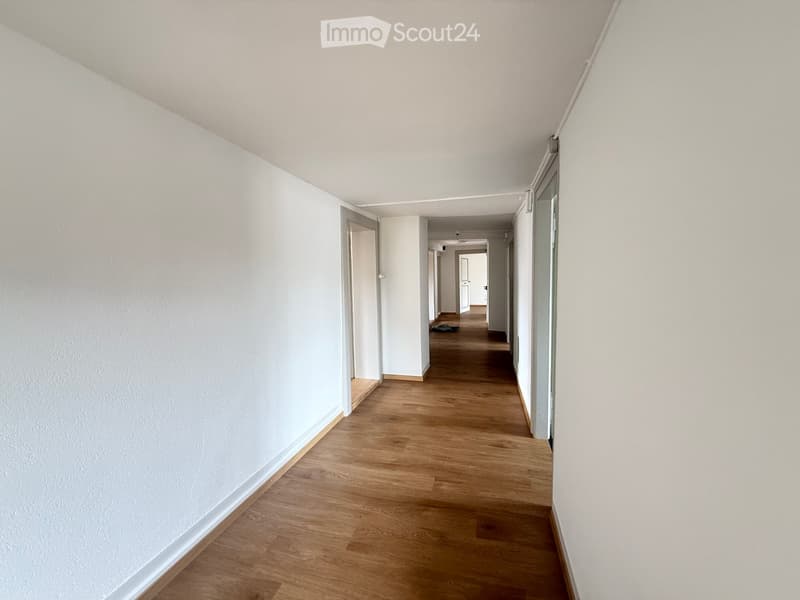 Empty hallway with light wooden flooring and white walls, featuring several doors and a distant view of another hallway.