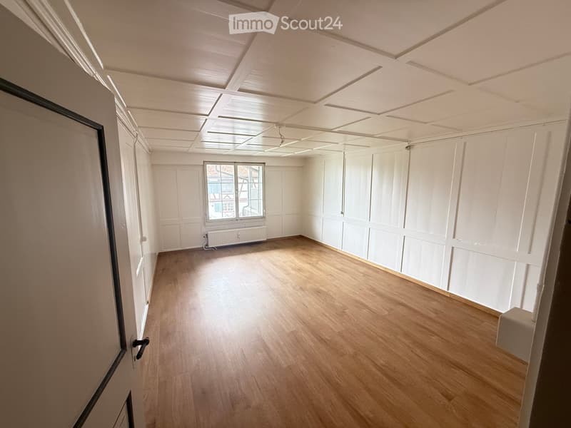 Empty room with wooden flooring, white walls, and a large window letting in natural light. The room features a radiator under the window and a door on the left side.
