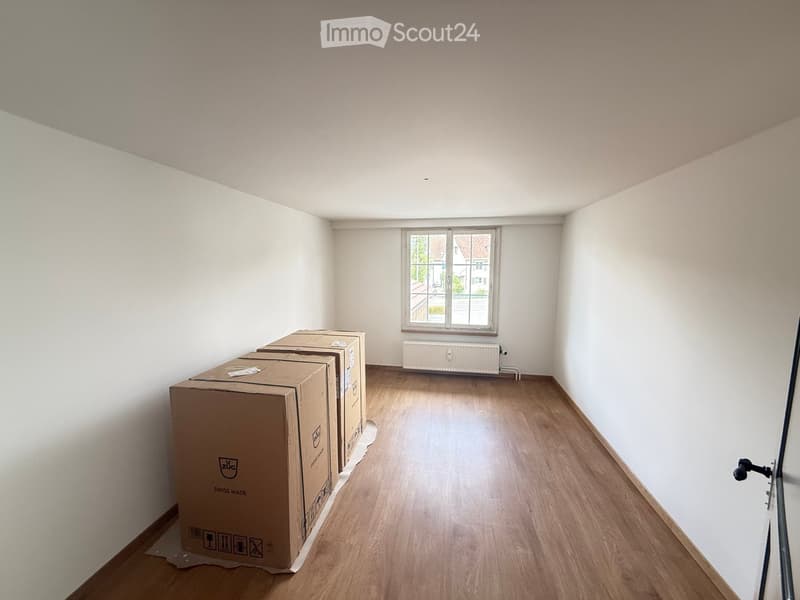 Empty room with light wooden flooring, white walls, and a window with a radiator below it. Several large cardboard boxes are stacked against the left wall.