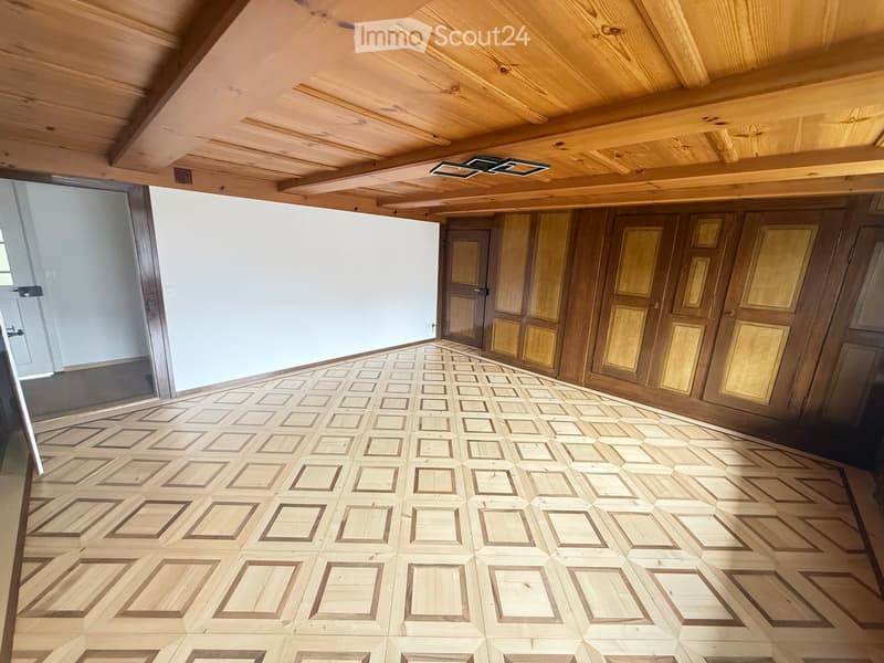 Empty room with wooden floor and exposed wooden ceiling beams. The walls are paneled with wood, and there are doors visible at the far end.