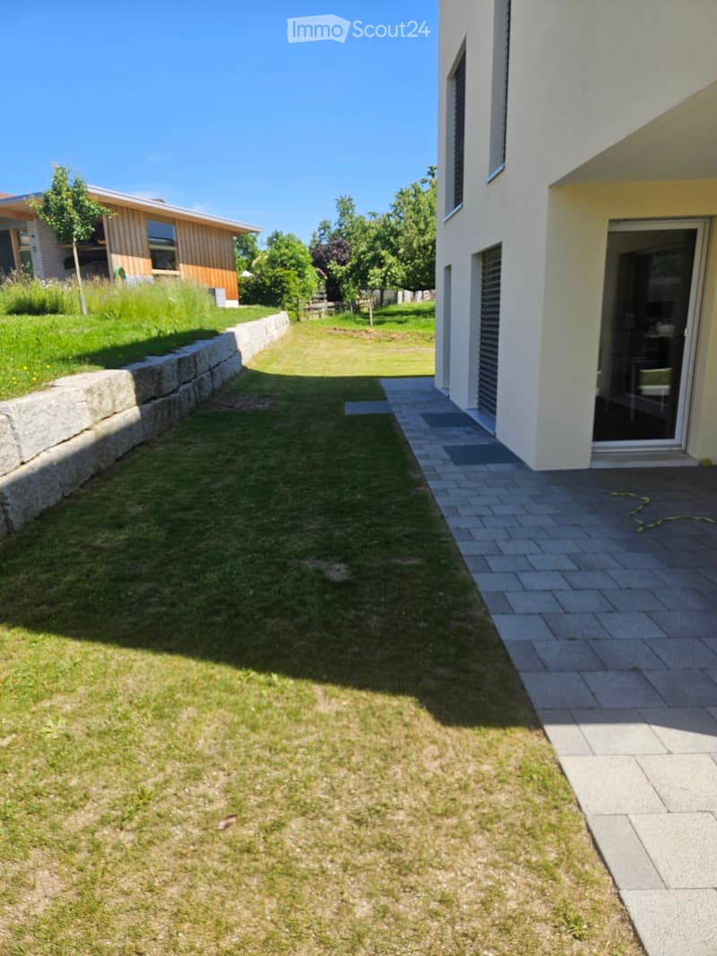 Well-maintained garden with a stone retaining wall, lawn, and paved pathway leading to a modern house entrance.