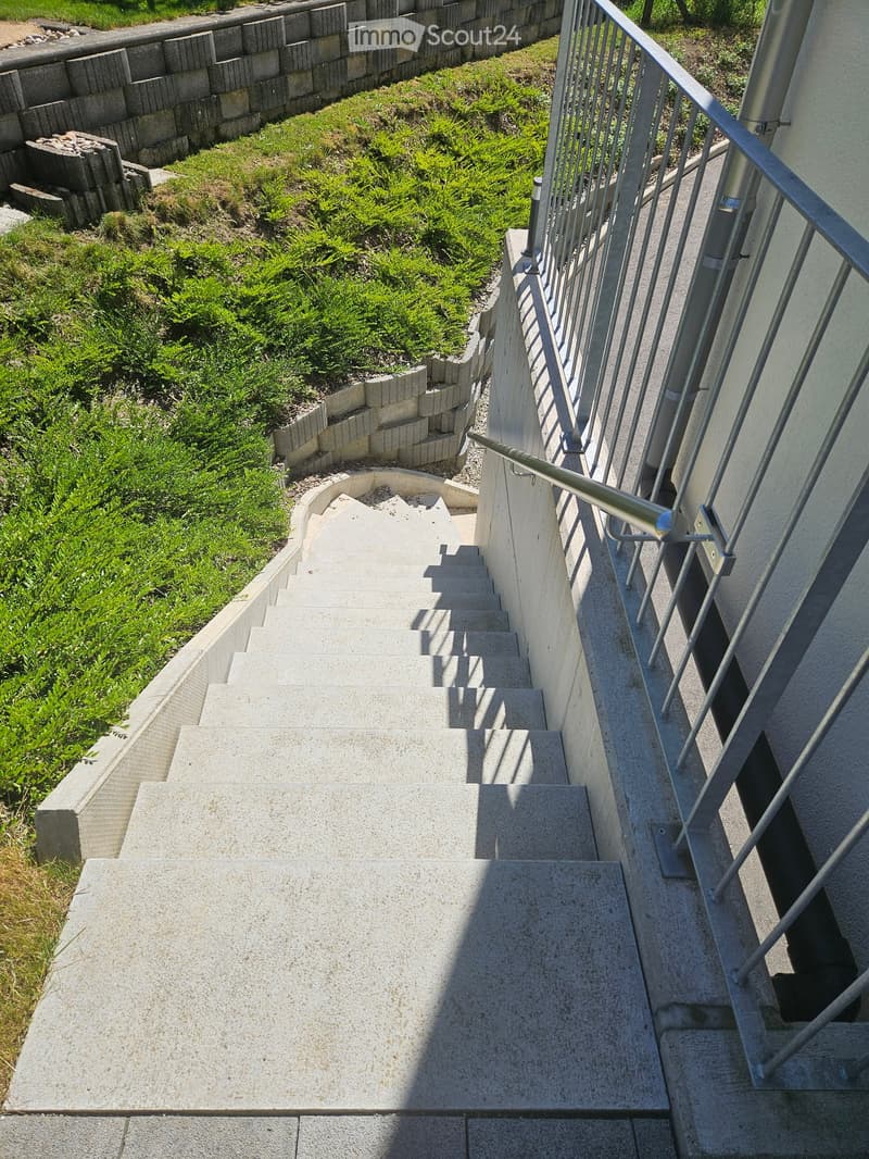 Concrete staircase with metal railing, leading up to a building entrance, surrounded by greenery and a low stone wall.