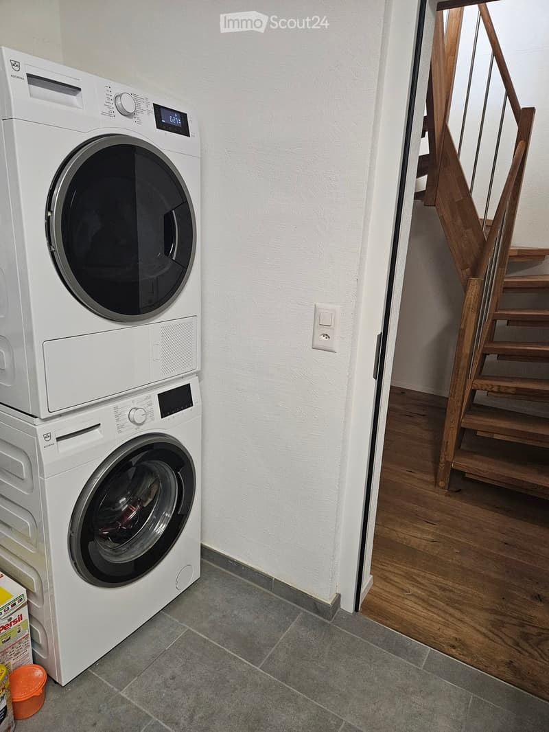 A small utility room with a washing machine and dryer stacked vertically, wooden staircase leading upwards, tiled floor, and white walls.