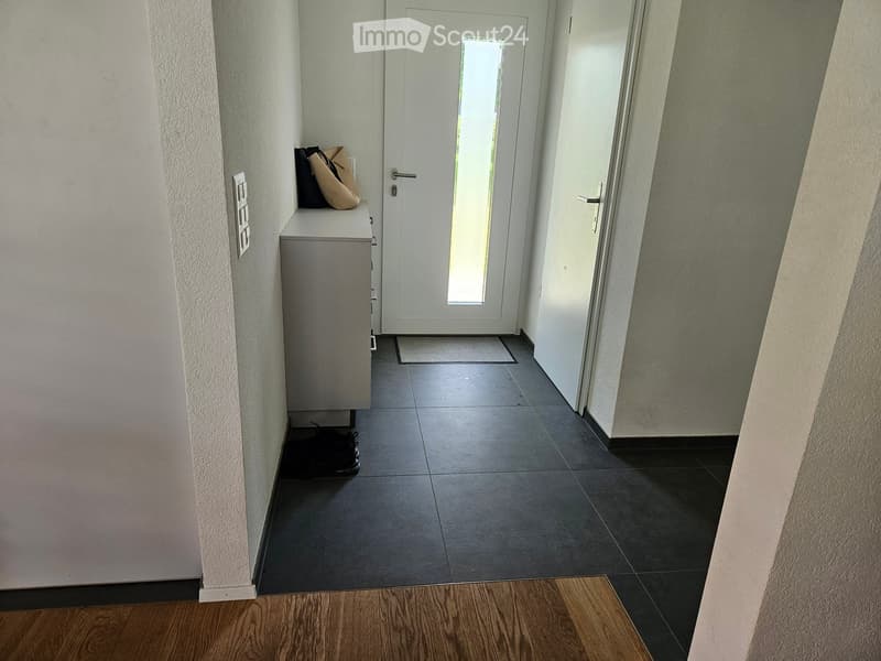 A narrow hallway with a tiled floor, white walls, and a white door with a glass panel at the end. A small cabinet with a bag on top is visible on the left side.