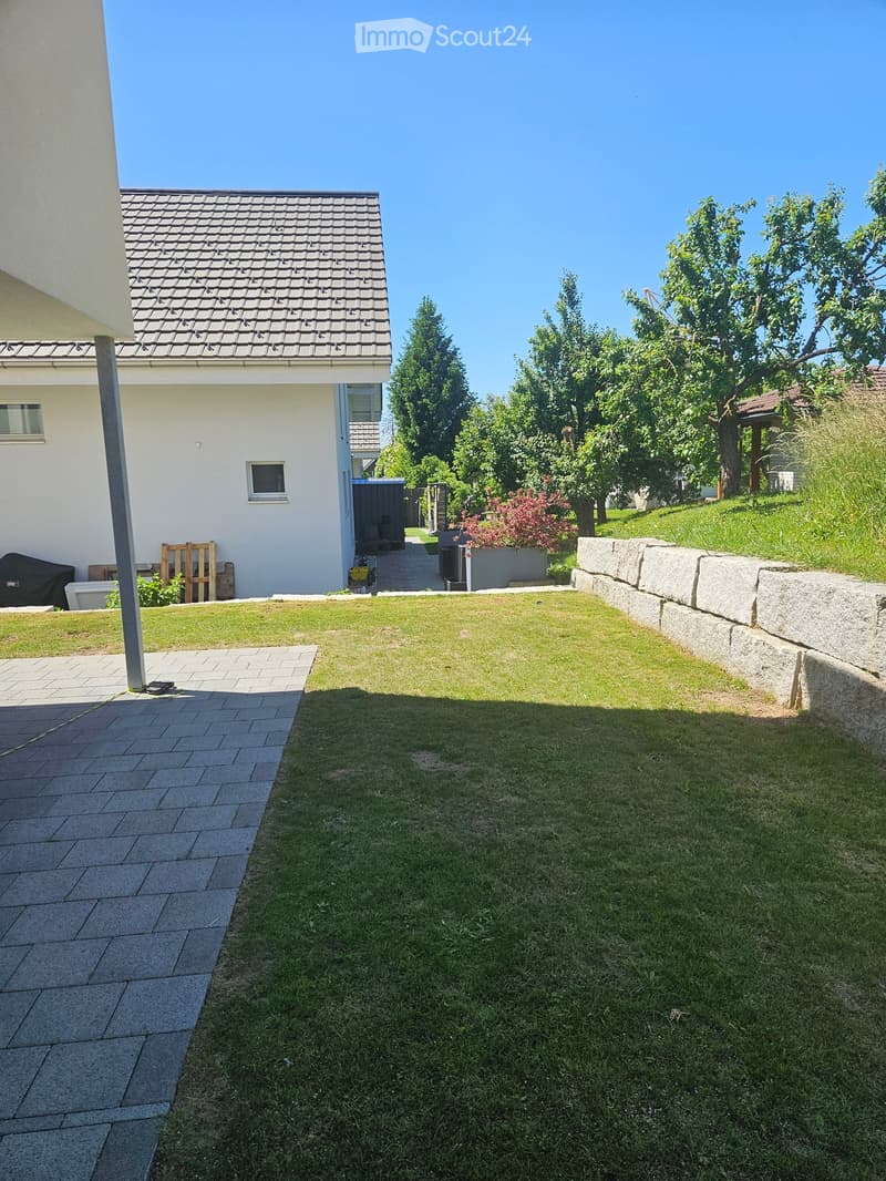 Well-maintained garden with a lawn, paved pathways, and a stone wall. The house is visible in the background with a covered patio area and some outdoor furniture.