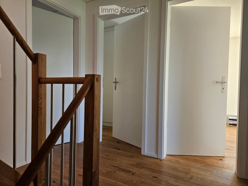 A modern hallway with wooden flooring, white walls, and a staircase with a wooden handrail and metal balusters. Two closed white doors are visible, one leading to another room.