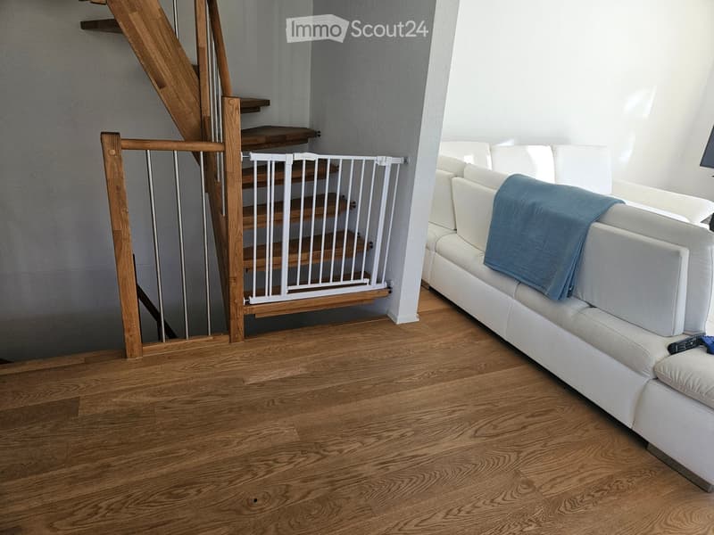 A modern living room with a white leather sofa, a blue blanket, a remote control, and a wooden staircase with a safety gate.