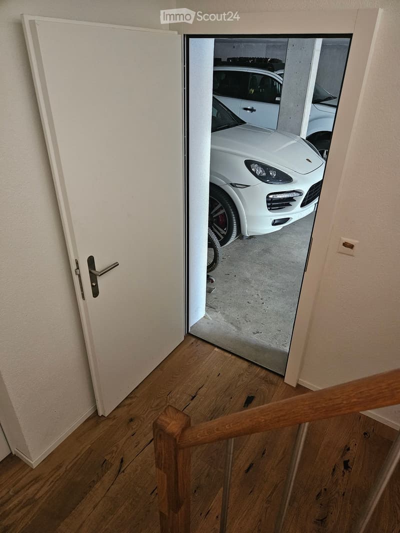 A wooden staircase leads to a doorway that opens into a garage with a white car parked inside.