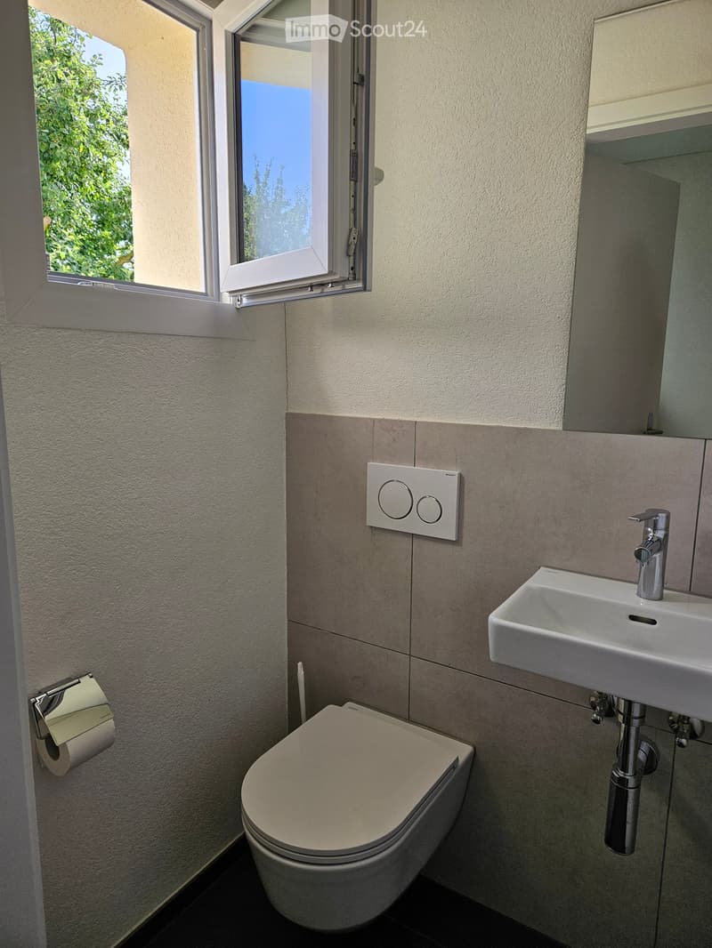 Modern bathroom with a toilet, sink, and faucet. Light-colored tiles and a window with blinds are visible.