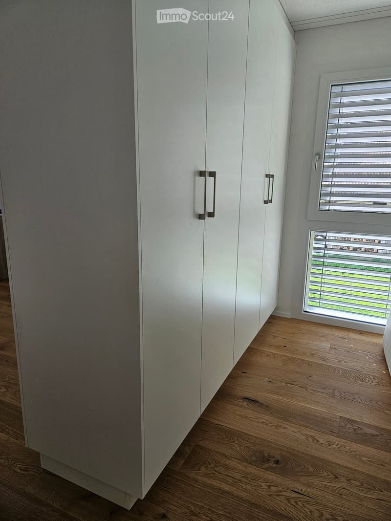 Empty room with light wooden flooring and white built-in wardrobes, featuring two windows with blinds.