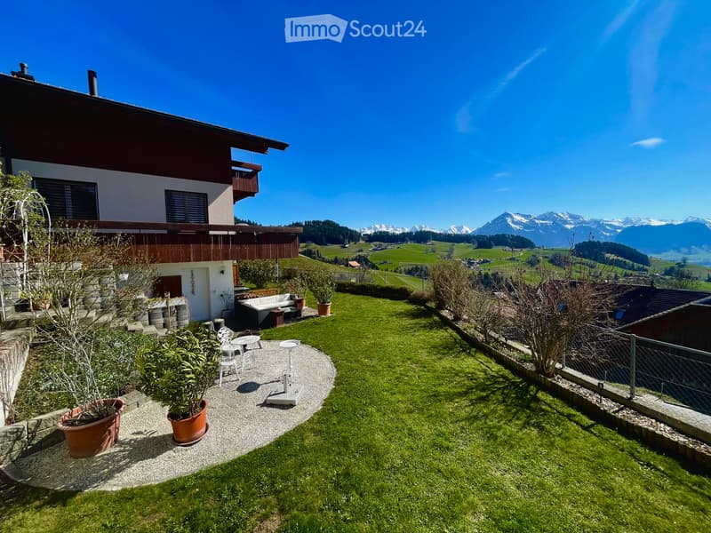 Garden / Outdoor Area. Garden with a well-maintained lawn, patio seating area with a sofa, table, and chairs, surrounded by potted plants and a scenic mountain view in the background.