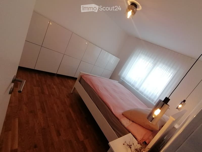 Bedroom with wooden floor, white walls, bed, white headboard, white nightstand, white cabinets, white curtains, ceiling lamps