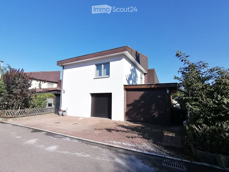 white house, brown roof, 2 garages, brick driveway, plants, trees