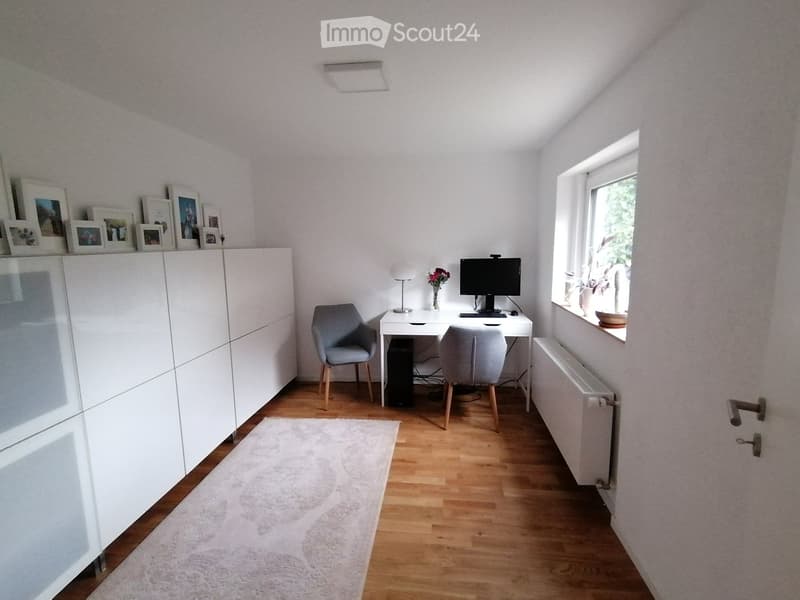 white walls, wooden floor, white desk, two chairs, computer monitor, lamp, radiator, rug, picture frames on the wall