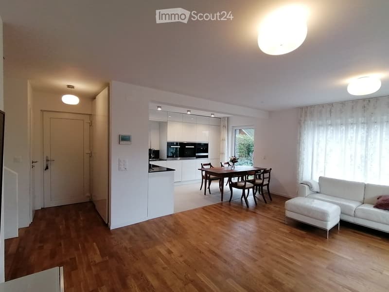 Open plan living and dining area with wooden floor, white walls, modern kitchen, dining table, white couch, and white ottoman