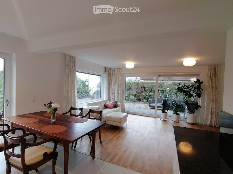 Dining room with wooden table, chairs, white couch, glass doors, plants, and a view of the outside